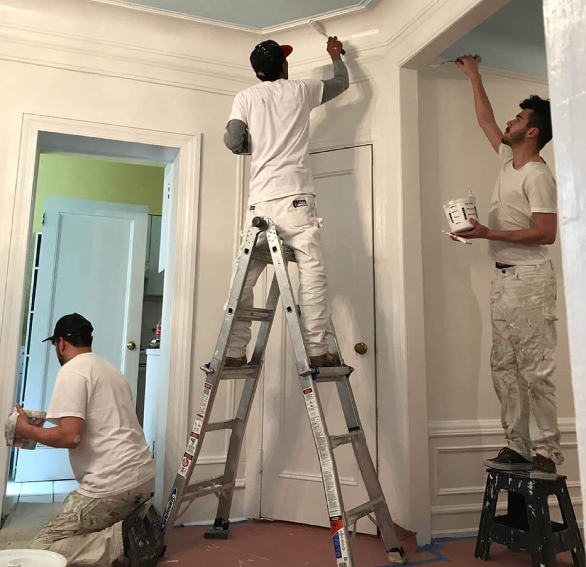 Two painters in a prewar apartment, one on a ladder cutting in along the door trim